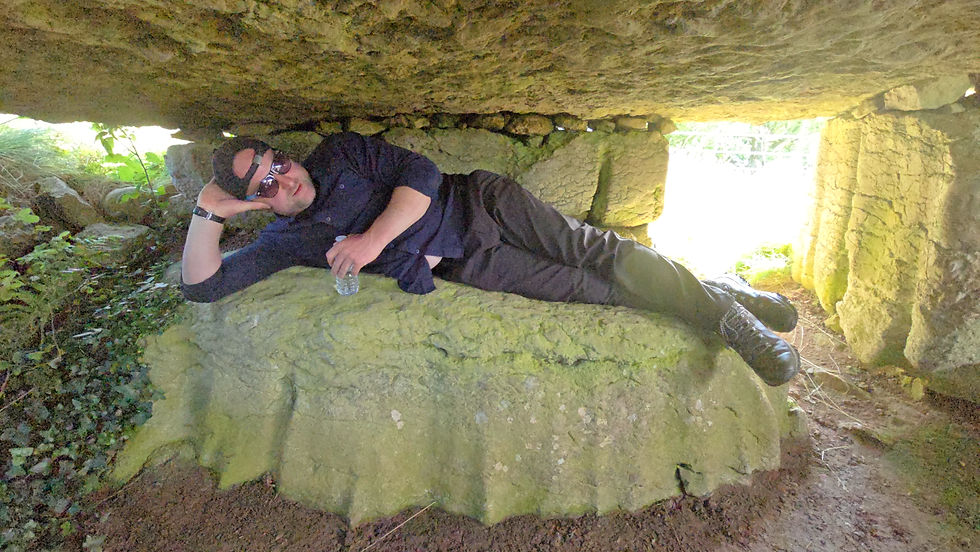 A man lying on the horizontal stone inside Lligwy Neolithic Burial Chamber beneath the low limestone capstone.