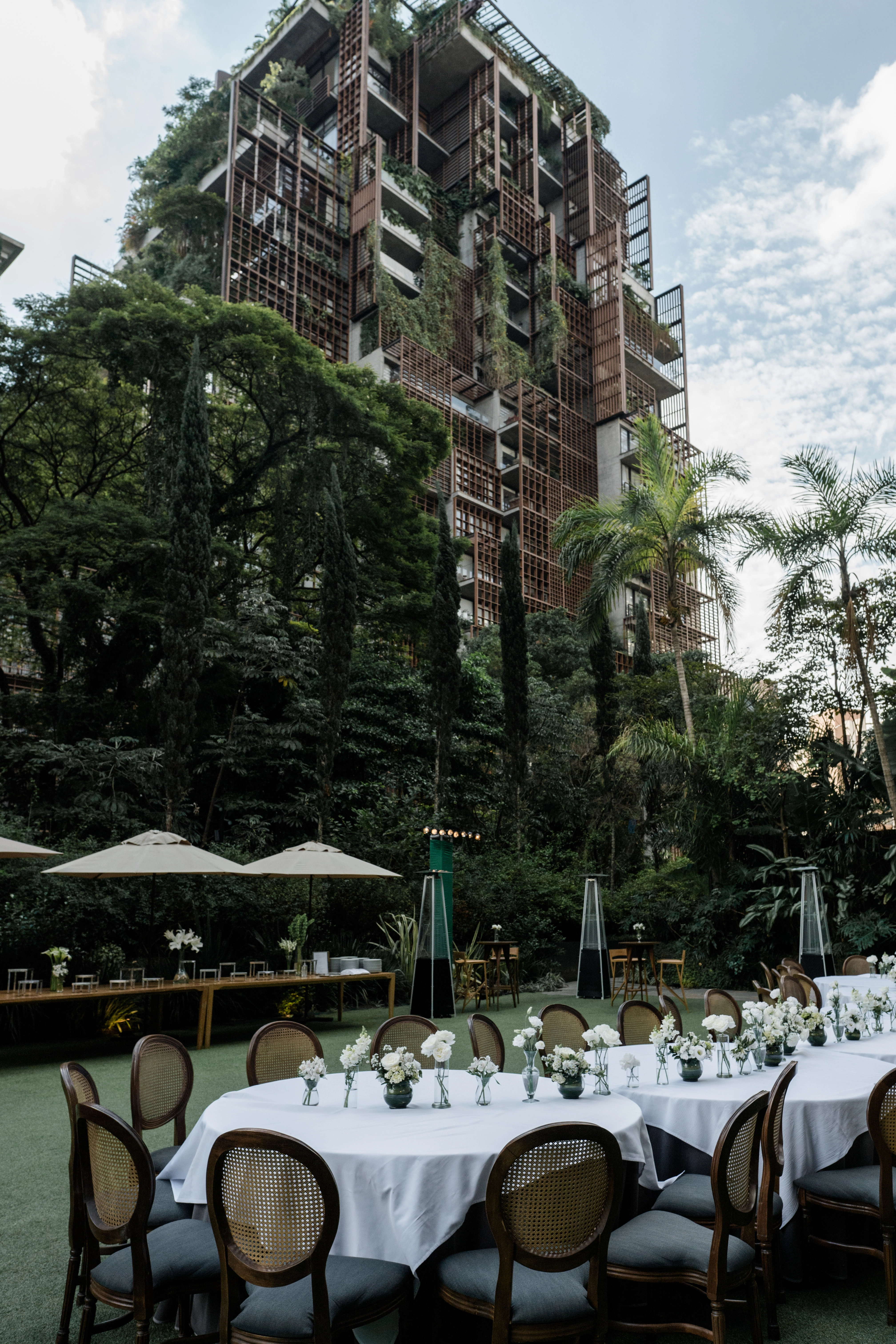 Close-up de mesas de casamento decoradas com pequenos vasos de flores brancas e cadeiras medalhão de palha sob iluminação natural do sol.