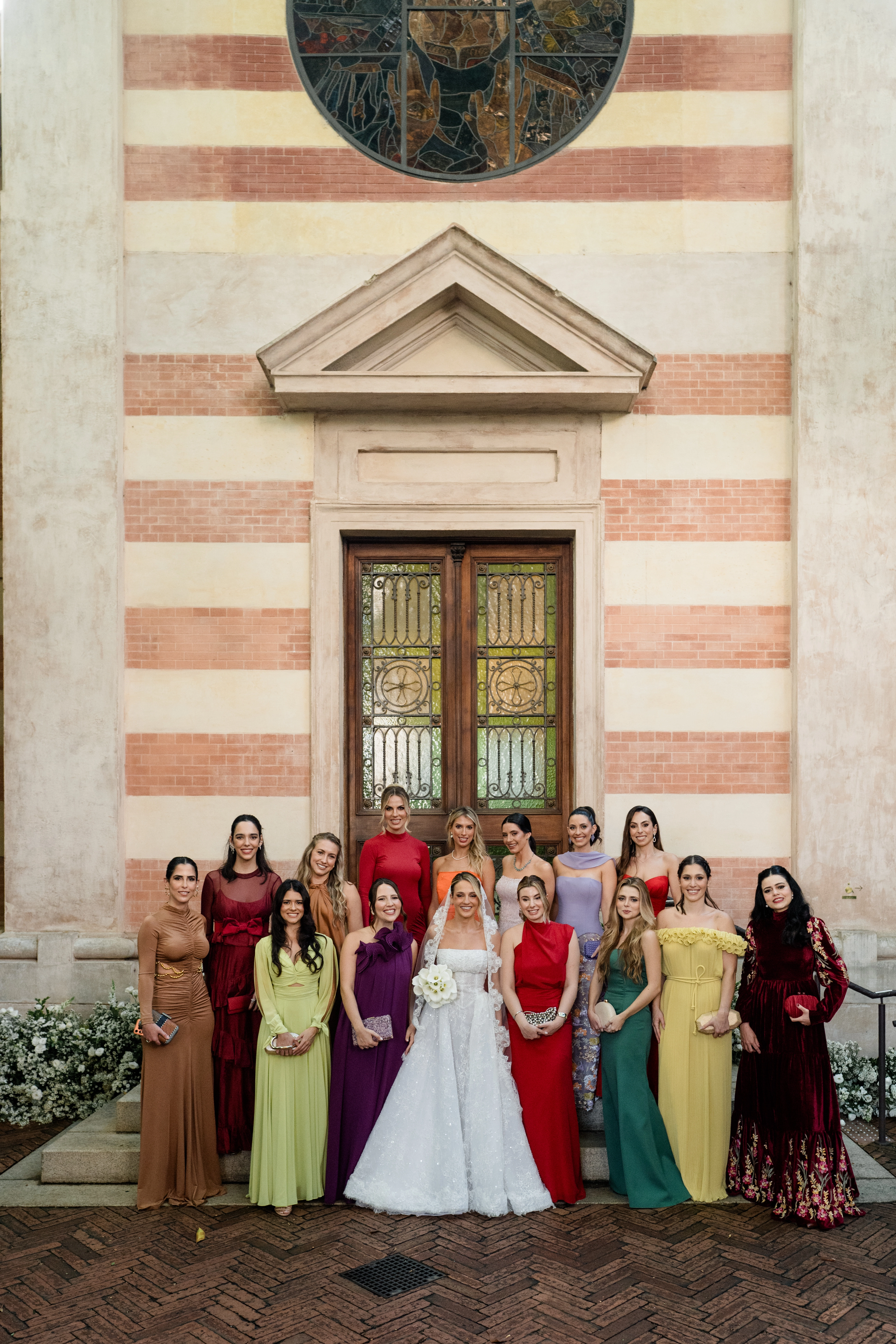 Noiva ao centro cercada por suas madrinhas vestindo vestidos longos em tons sofisticados de vermelho, roxo, verde e amarelo na escadaria externa da igreja.