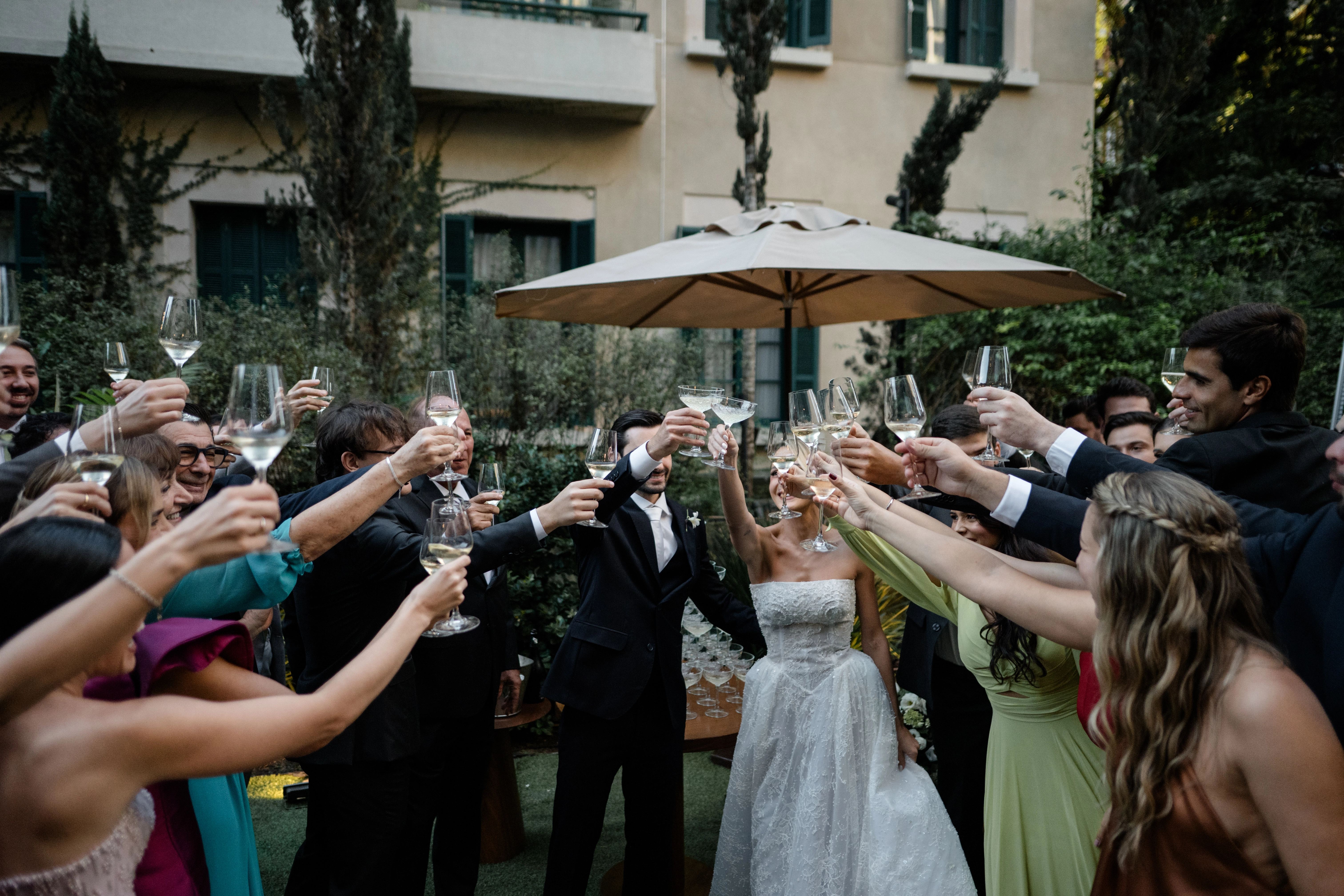 Noivos e convidados sorrindo e brindando com taças de champanhe em uma celebração de casamento sofisticada ao ar livre durante o dia.