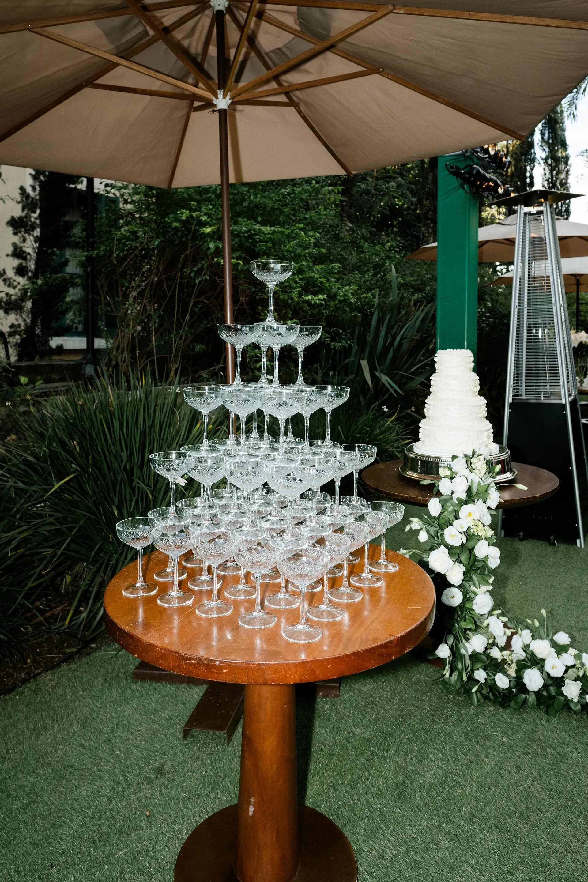 Torre de taças de champanhe de cristal lapidado montada sobre mesa redonda de madeira em recepção de casamento elegante ao ar livre.
