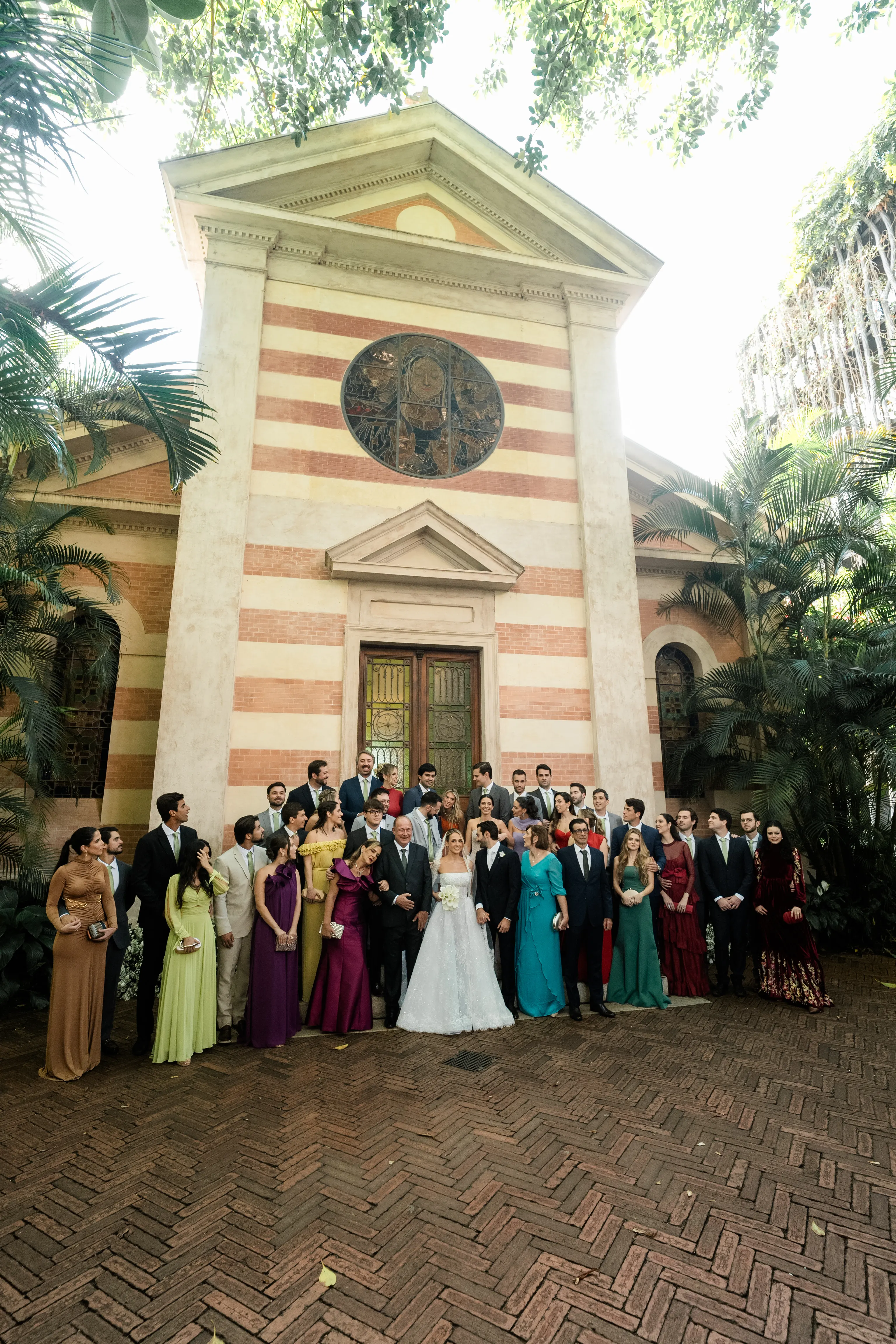 Imagem com plano aberto mostrando o grande grupo de convidados do casamento posando em frente à fachada monumental da igreja, emoldurada por palmeiras e plantas tropicais.