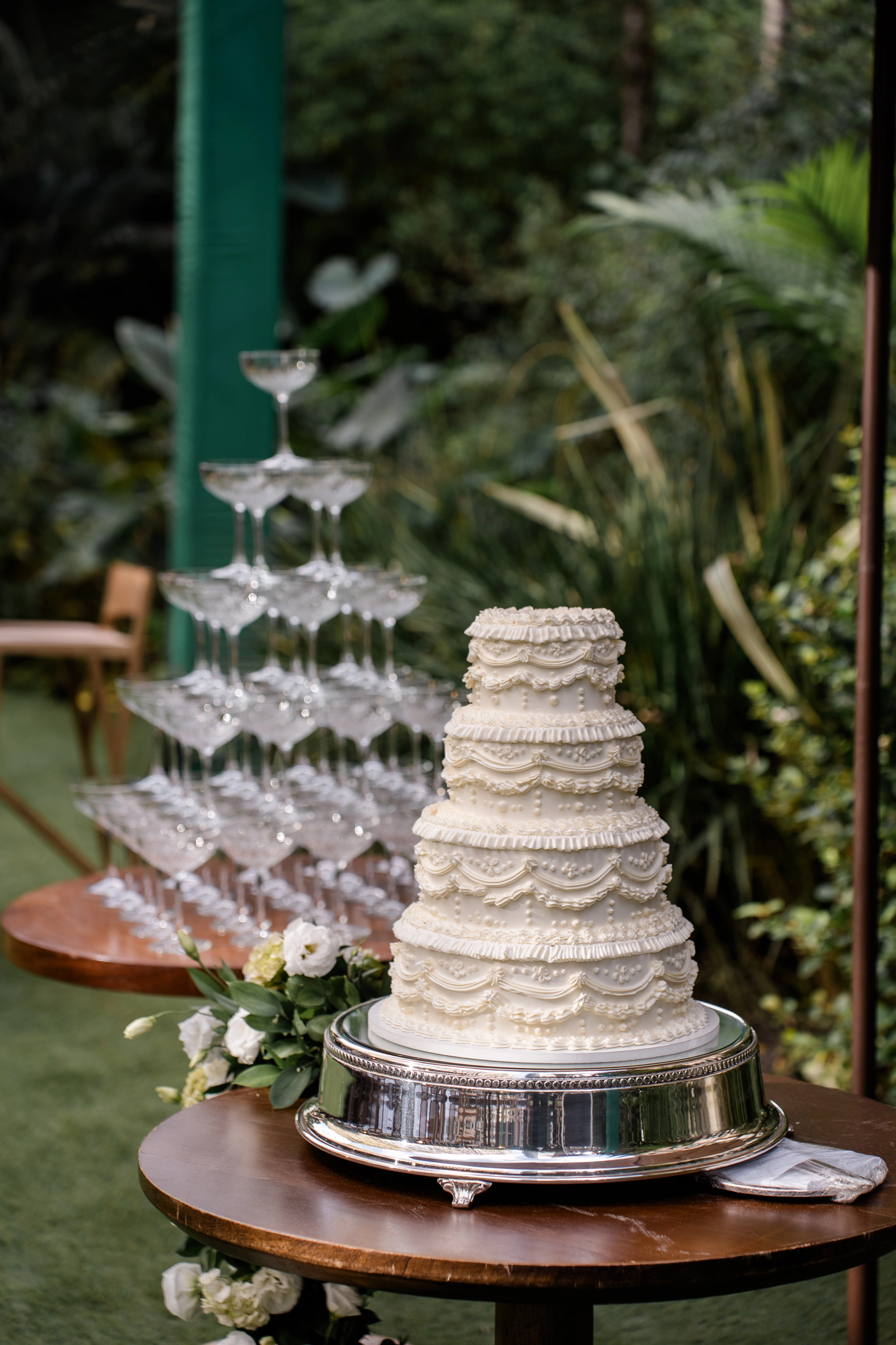 Bolo de casamento artesanal texturizado em primeiro plano com uma torre de champanhe desfocada ao fundo em ambiente externo sob ombrelone.