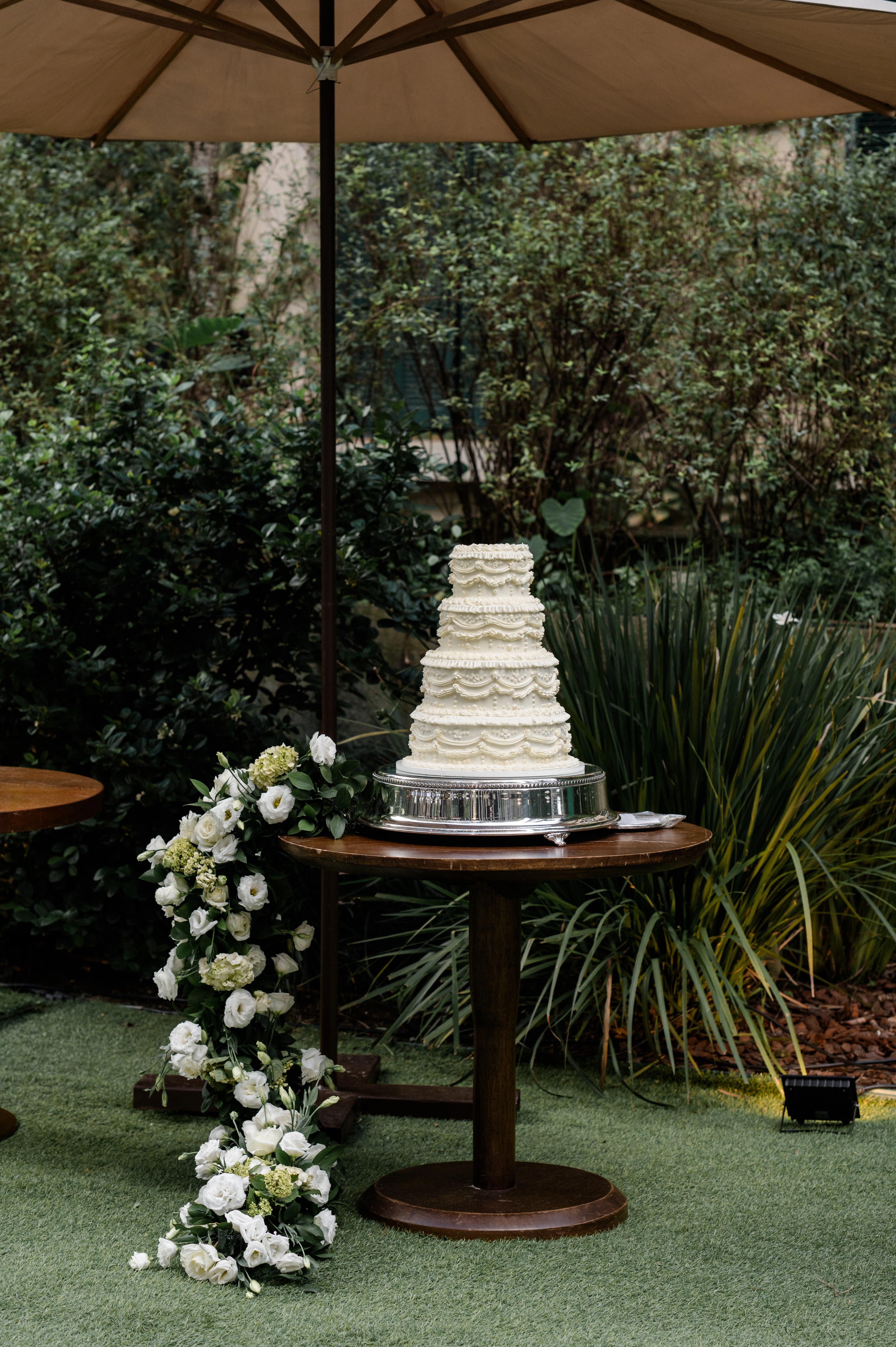 Bolo de casamento clássico de quatro andares com acabamento de glacê trabalhado, posicionado sobre mesa de madeira com arranjo de flores brancas ao ar livre.