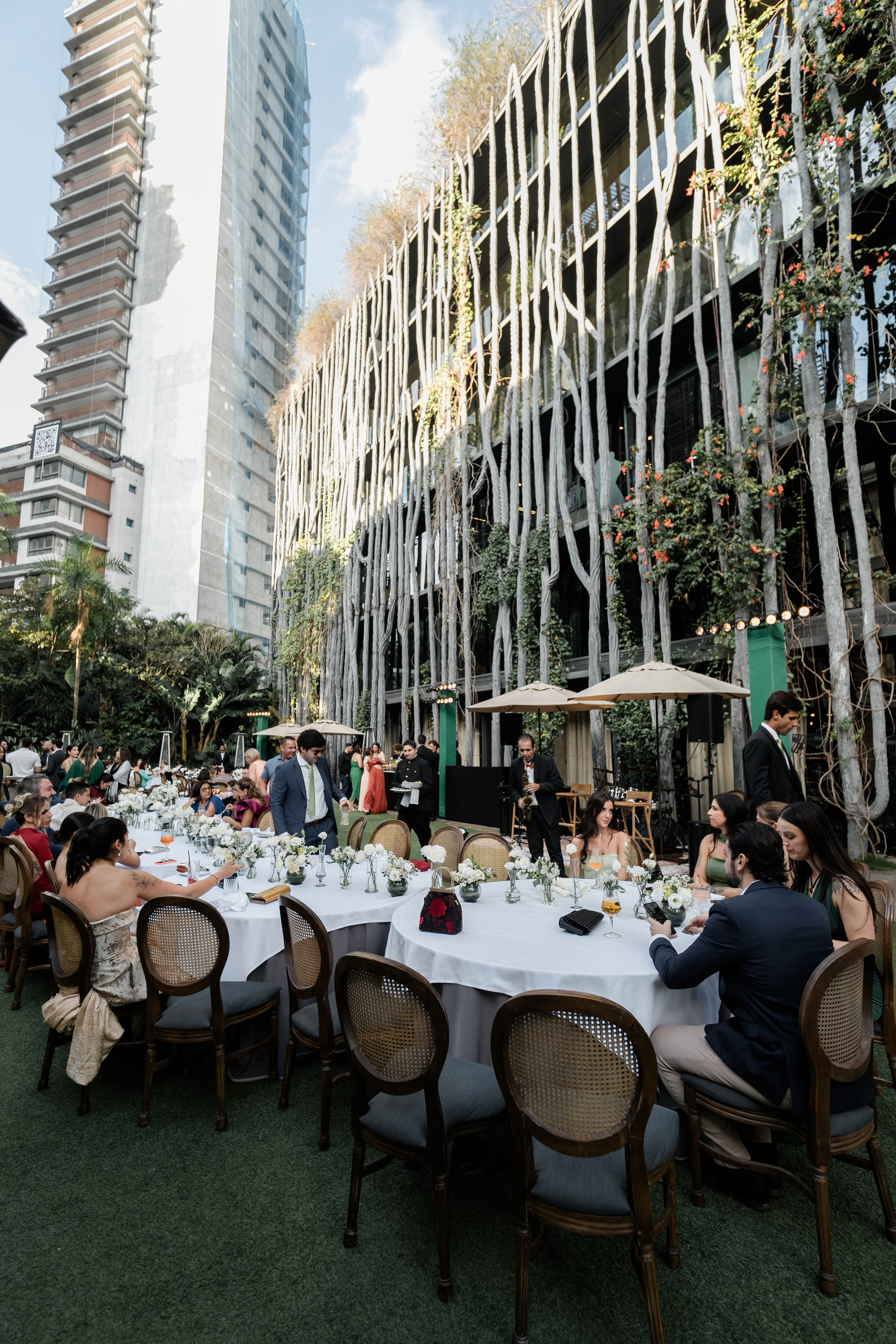 Convidados assentados em mesas longas de casamento ao ar livre, conversando sob ombrelones em um jardim de luxo.