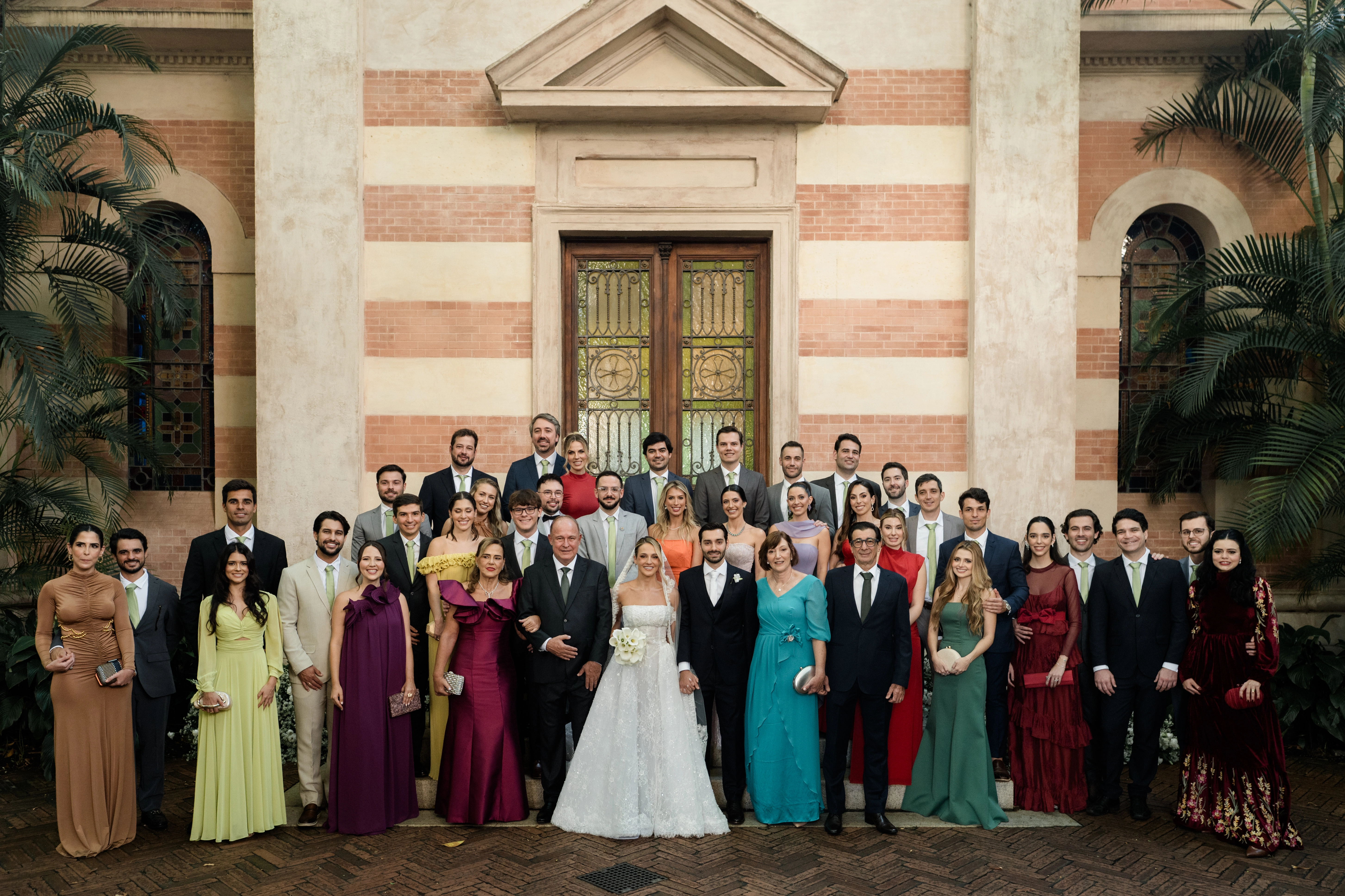 Retrato oficial de casamento com noivos, padrinhos e madrinhas posando organizados em frente à fachada de tijolos listrados de uma igreja clássica.