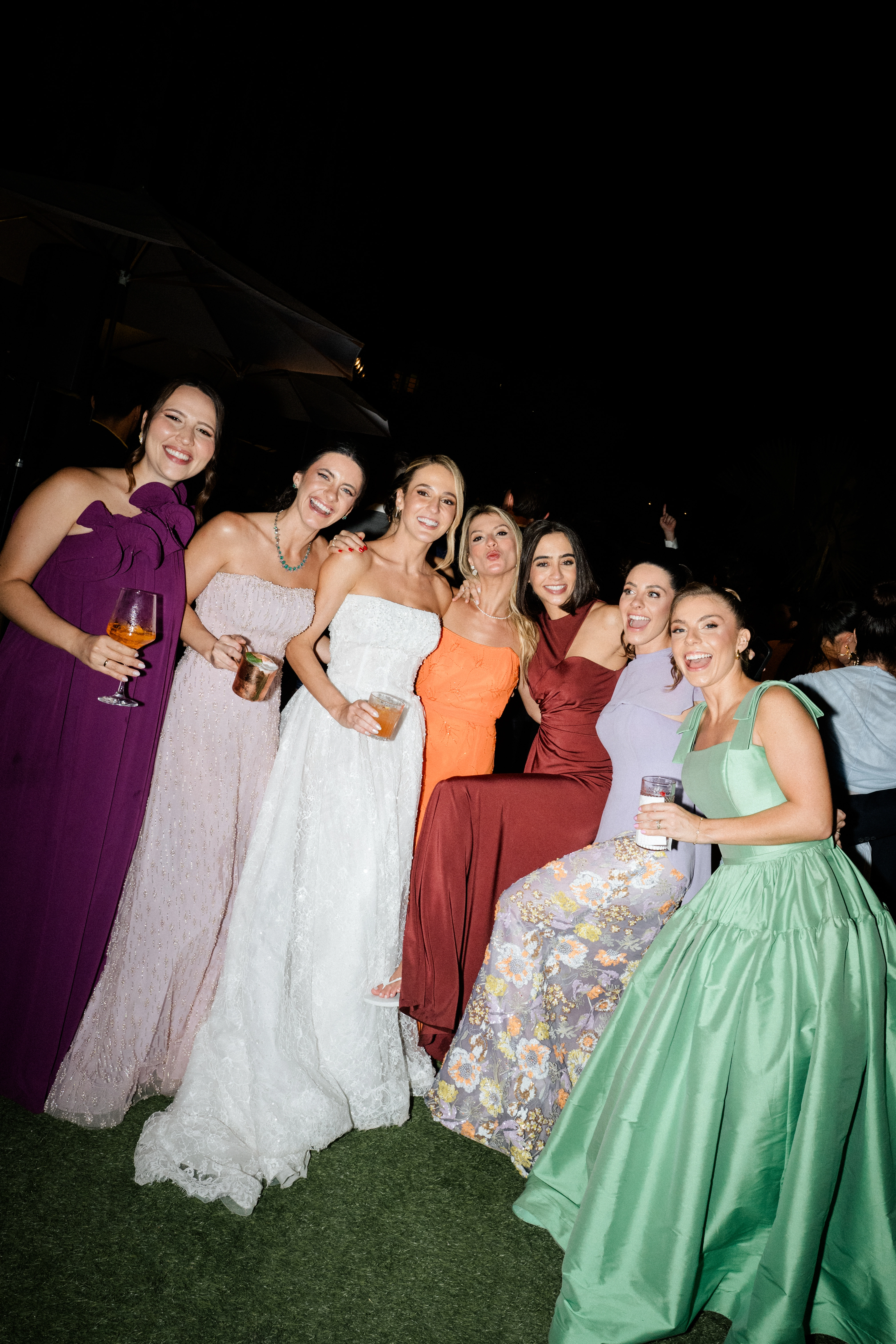 Noiva sorridente ao lado de suas amigas e madrinhas segurando taças de drinks em uma recepção de casamento de luxo ao ar livre durante a noite.