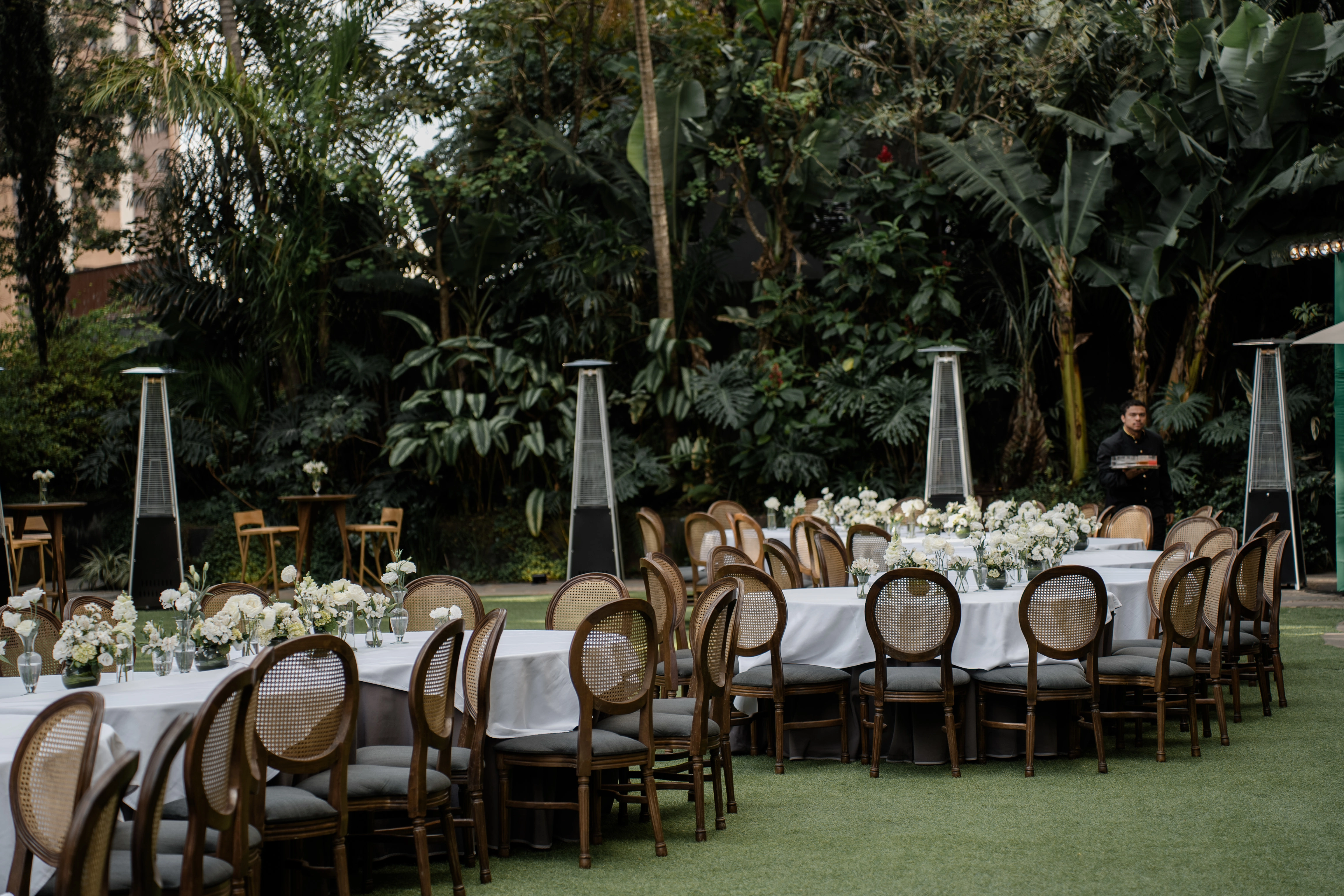 Decoração de casamento de luxo ao ar livre com mesas redondas de toalhas brancas, cadeiras de palha clássicas e arranjos florais minimalistas sobre o gramado.