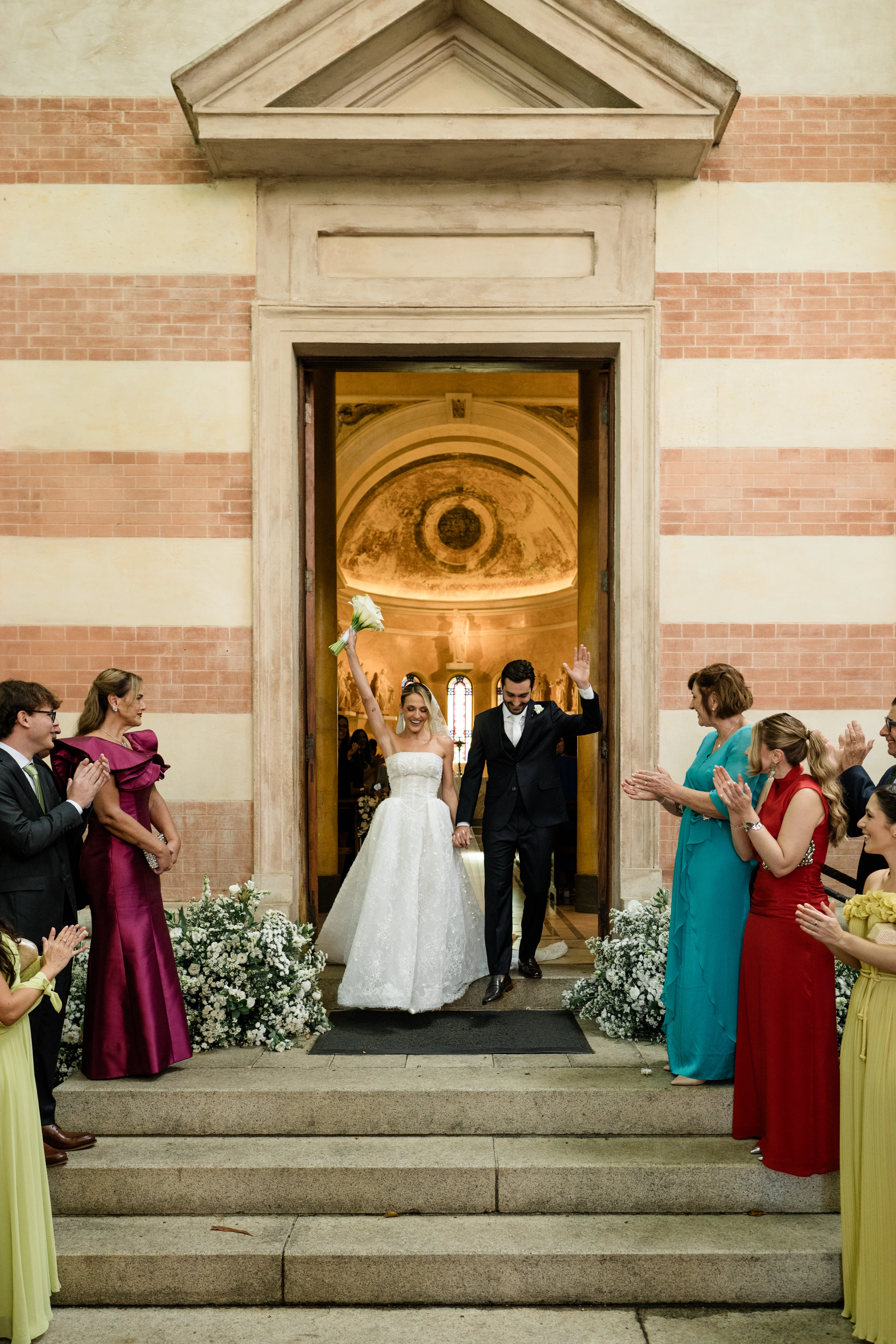 Noivos recém-casados saindo pelas portas de madeira da igreja de mãos dadas, sob os aplausos e sorrisos dos padrinhos nos degraus de pedra.