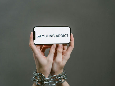 Chained hands hold a phone displaying the text 'Gambling Addict', highlighting addiction.