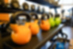 Orange and green kettlebells on a gym rack, labeled "25," with dumbbells on the shelf above. Bright, spacious gym setting.