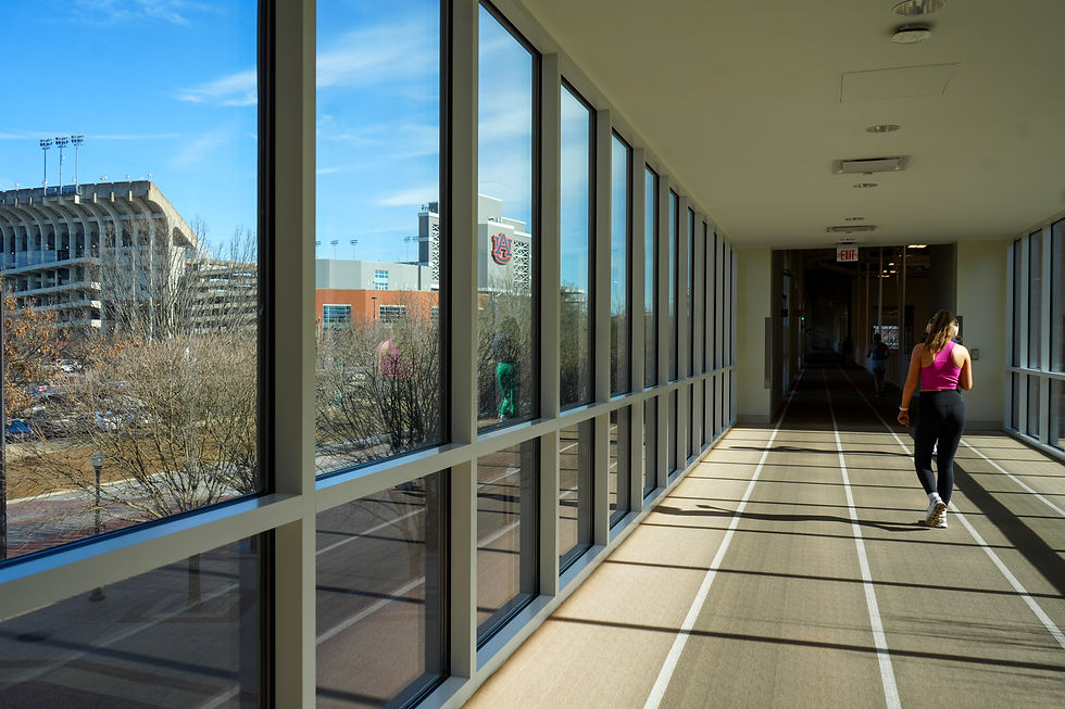 A woman in a pink top walks on an indoor track. Large windows show a stadium outside. Bright daylight and shadows enhance the modern setting.