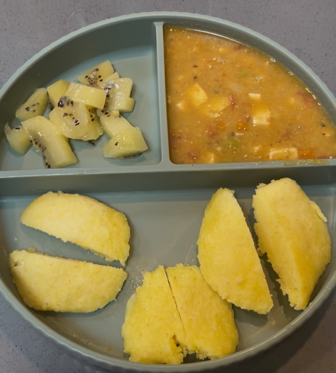 Moong dal idli cut into pieces served on a divided silicone plate with sambar and kiwi fruit — complete baby meal idea India starting solids