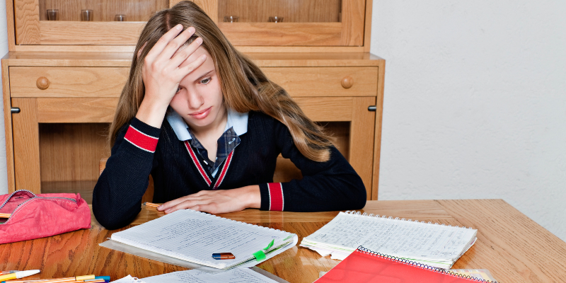 Girl in school uniform, appearing frustrated, holds head while studying at a wooden table with notebooks and pens. Background has wooden shelves.
