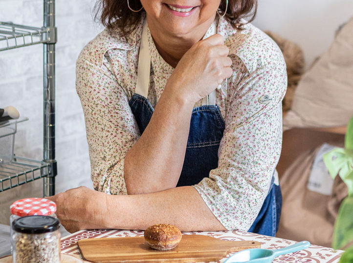 Mulher de pé na cozinha apoiada na bancada olhando os ingredientes e pães prontos.