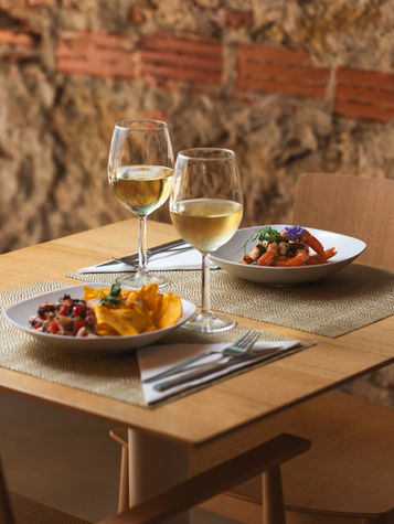 Mesa posta em um restaurante, com duas cadeias, dois pratos de comida e bebidas em cima da mesa. Ambiente acolhedor em tons marrons e texturas em pedra na parede.