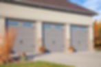 Three gray garage doors with stone accents, flanked by potted flowers. Sunny day, autumn trees in background, and tall grass in foreground.