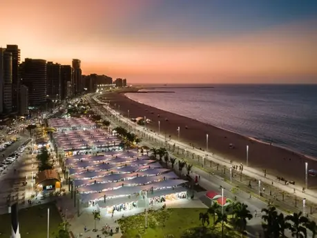 Vista da orla da Praia do Meireles ao pôr do sol. Na foto, é possível ver o amplo calçadão, a tradicional feirinha da beira mar e um misto de prédios e do mar.