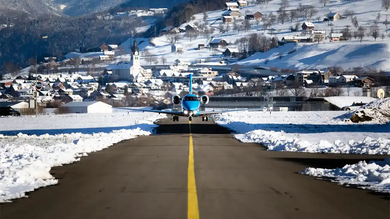 Blue private jet pilatus pc-24 on a snowy runway, with a mountain village and white church in the background.