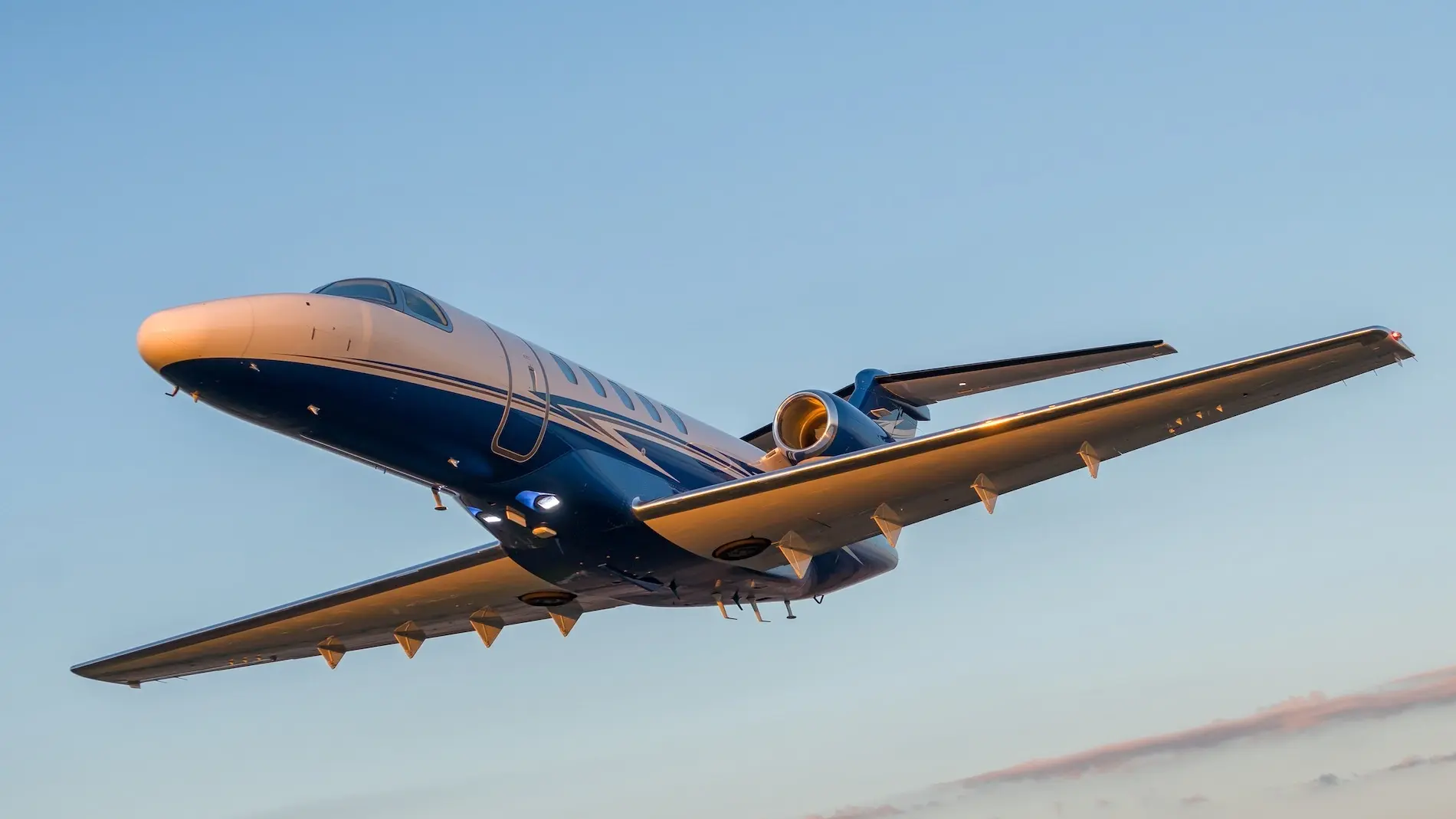 Citation CJ4 private jet flying low against a clear blue sky at sunset, with soft pink clouds below.