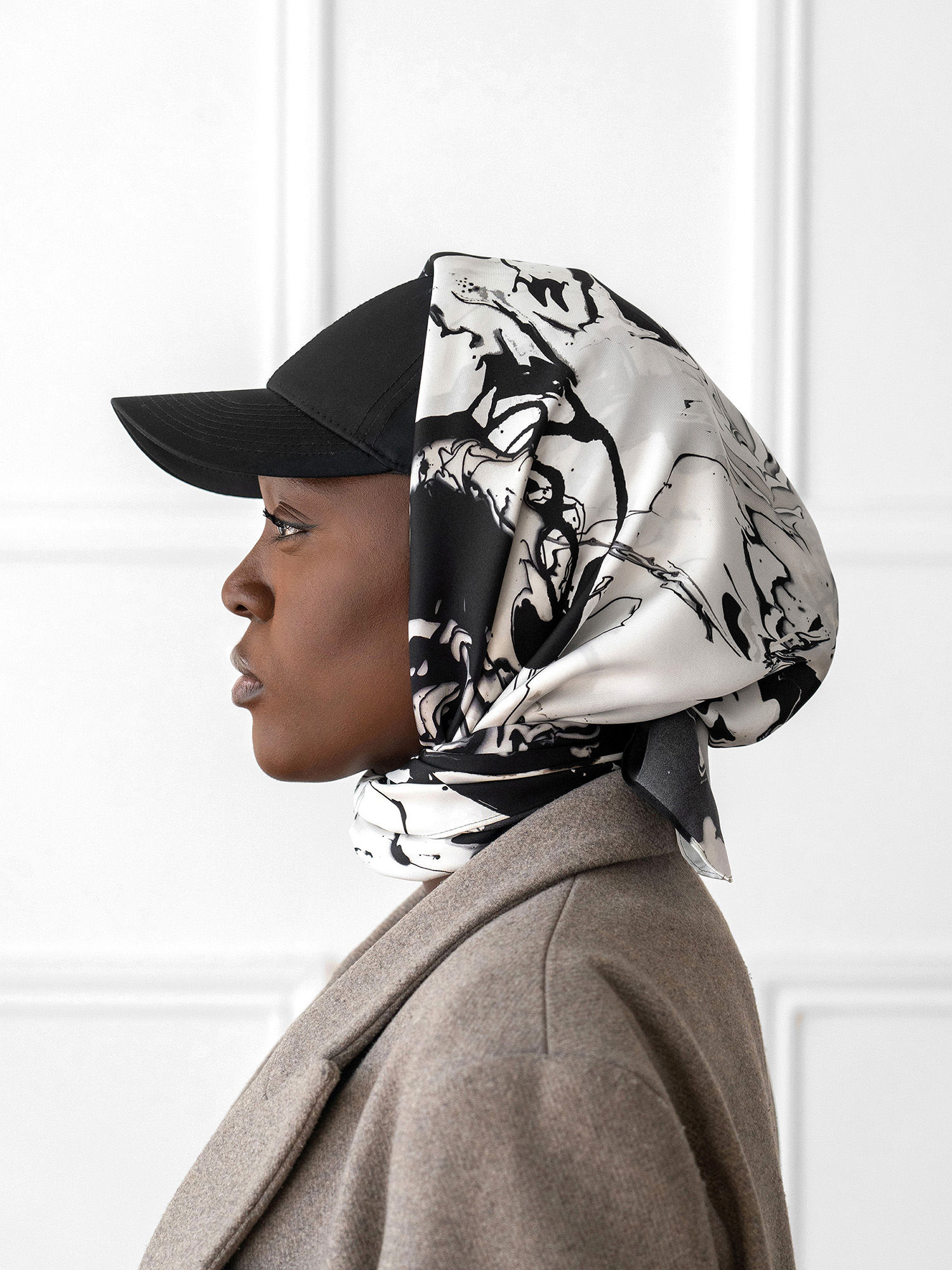 A profile view of a model in brown coat with a black and white patterned silk scarf over her head.