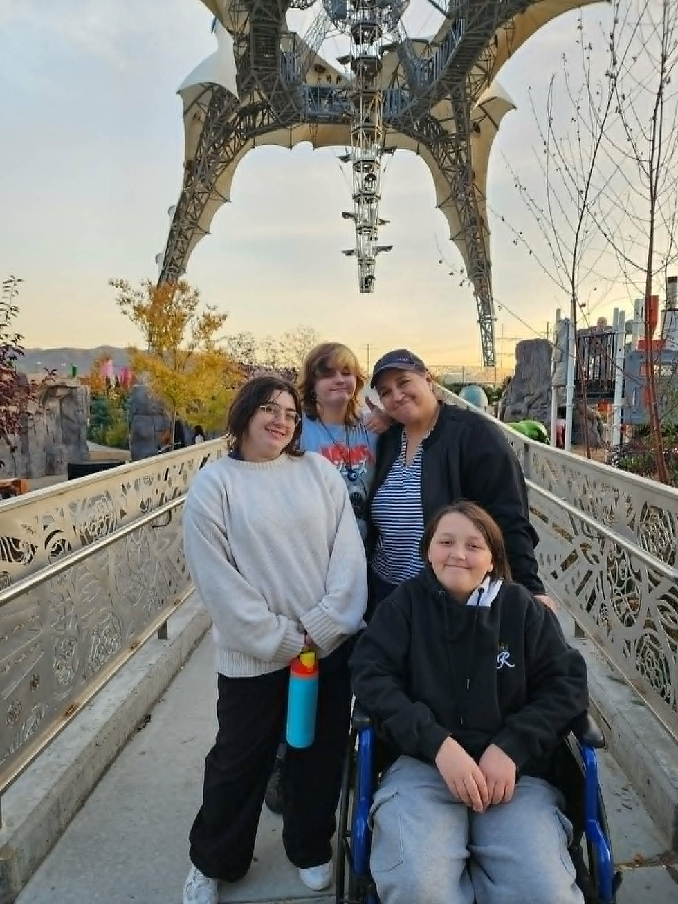 A family standing on a walkway outside. A boy in a wheelchair in front with 3 other family members behind. There is a light and sound system above their heads. Its is winter time. The trees are loosing leaves or have none.