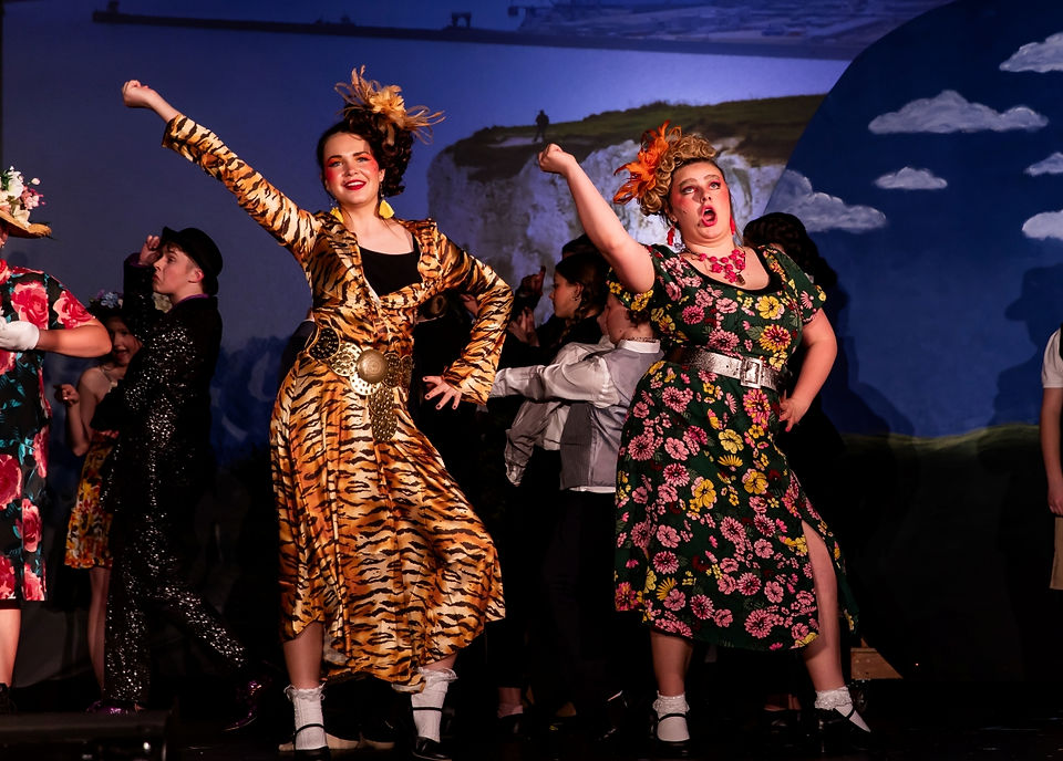 Two young actors, dressed in vibrant costumes—a tiger-striped dress and a floral dress with dramatic accessories—perform as Aunt Spiker and Aunt Sponge during James and the Giant Peach. They sing and dance with exaggerated expressions, bringing comedic energy to the scene.
