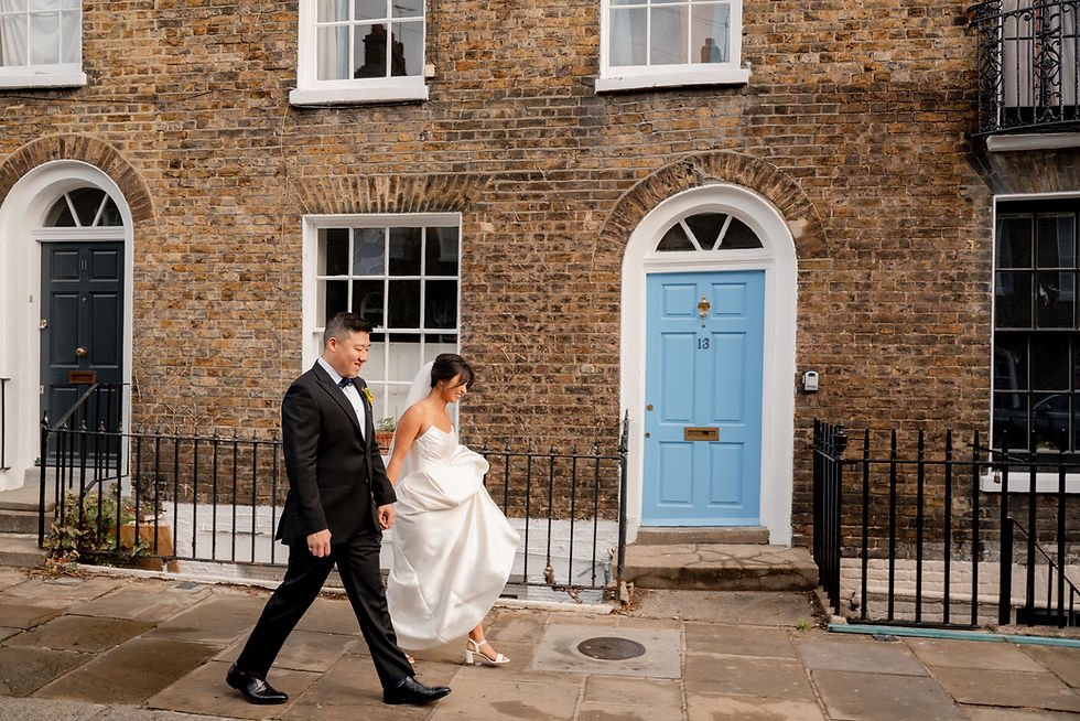 Bride and groom in formal attire walk past brick buildings with blue and black doors. Bride smiles, holding her dress. Door marked 13.