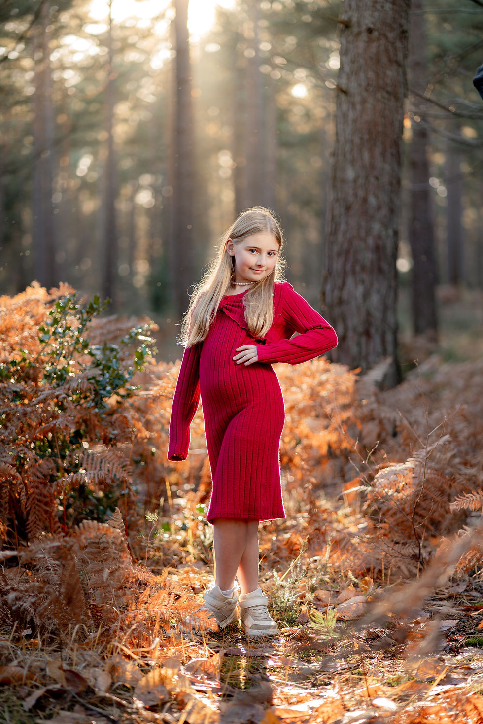 Girl in a red dress stands confidently in a sunlit forest, surrounded by vibrant brown ferns. Soft, golden light creates a warm mood.
