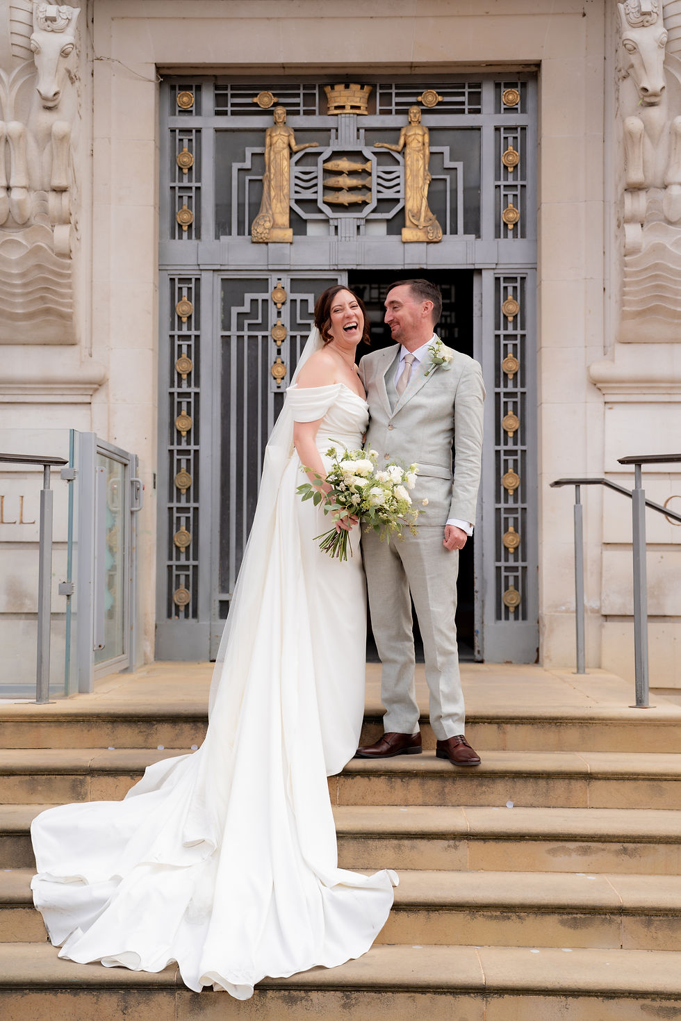 Bride and groom laugh on stone steps before an ornate Art Deco doorway, the bride in a white gown holding a bouquet.