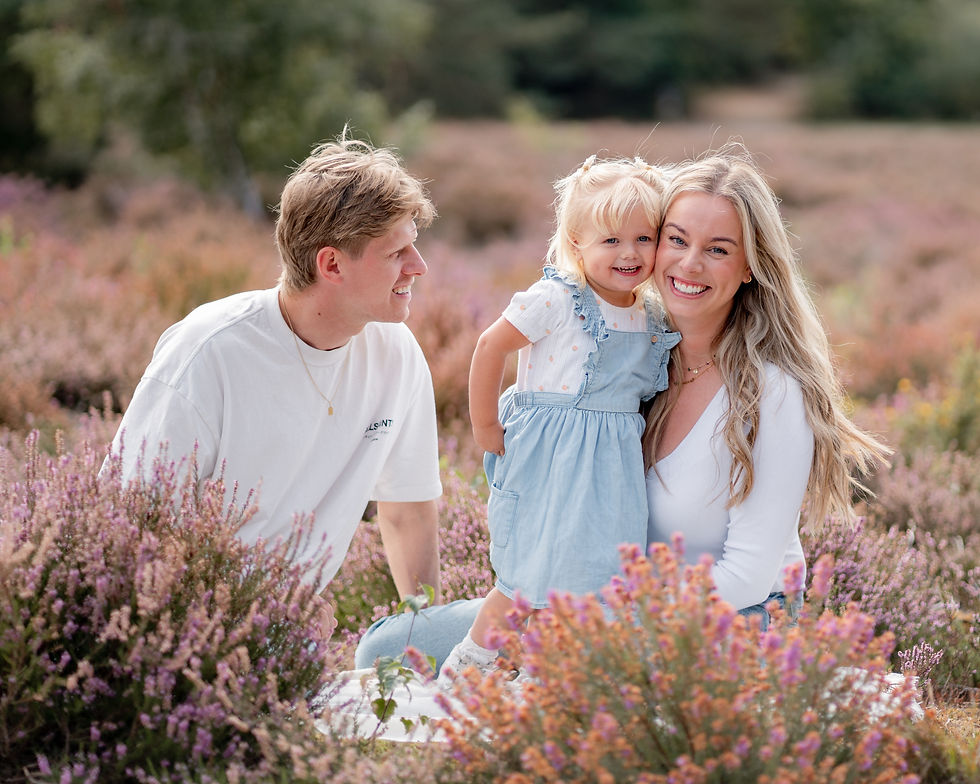 A smiling family sits in a field of purple flowers. The woman holds a young child in a blue dress. The man looks on, wearing a white shirt.