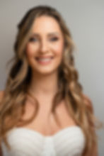 Smiling woman with wavy brown hair in a pearl-adorned white top against a simple gray background. Bright, happy expression.