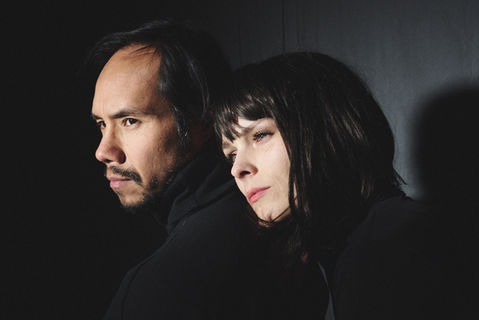Moody portrait of a man and a woman from the band Jurij Mondaine. They are positioned close together, both looking off-camera in profile. The lighting is dramatic, with bright highlights on their faces and deep shadows in the background. Both are wearing dark clothing, creating a minimalist and atmospheric synth-pop aesthetic. Captured in Hamburg by Félix L. Salazar