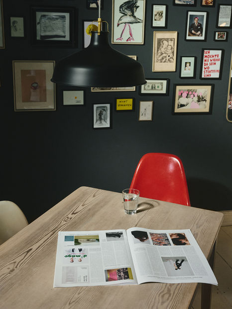 Open magazine and glass of water under a black pendant lamp, against a dark wall filled with frames.