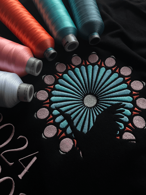 Broderie colorée d’une rosace stylisée entourée de bobines de fils sur tissu noir.