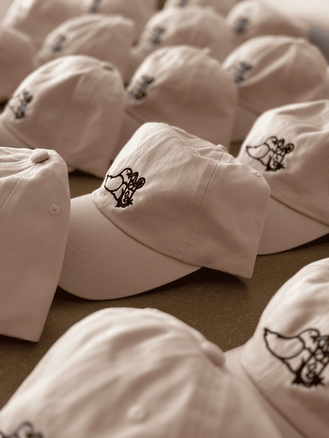 Casquettes beige brodées d’un logo cartoon noir, alignées en série.