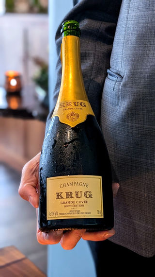 At Aubergine, a person is holding a bottle of Krug champagne, angled slightly for display. The iconic gold label is clearly visible, set against the backdrop of a refined dining space.