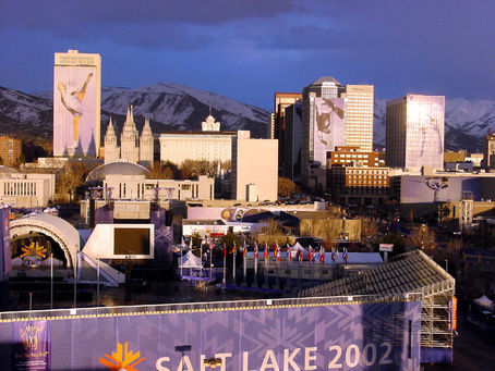 Salt Lake City skyline during 2002 Olympics, with skyscrapers, mountains, flags, and banners reading "Salt Lake 2002" at sunset.