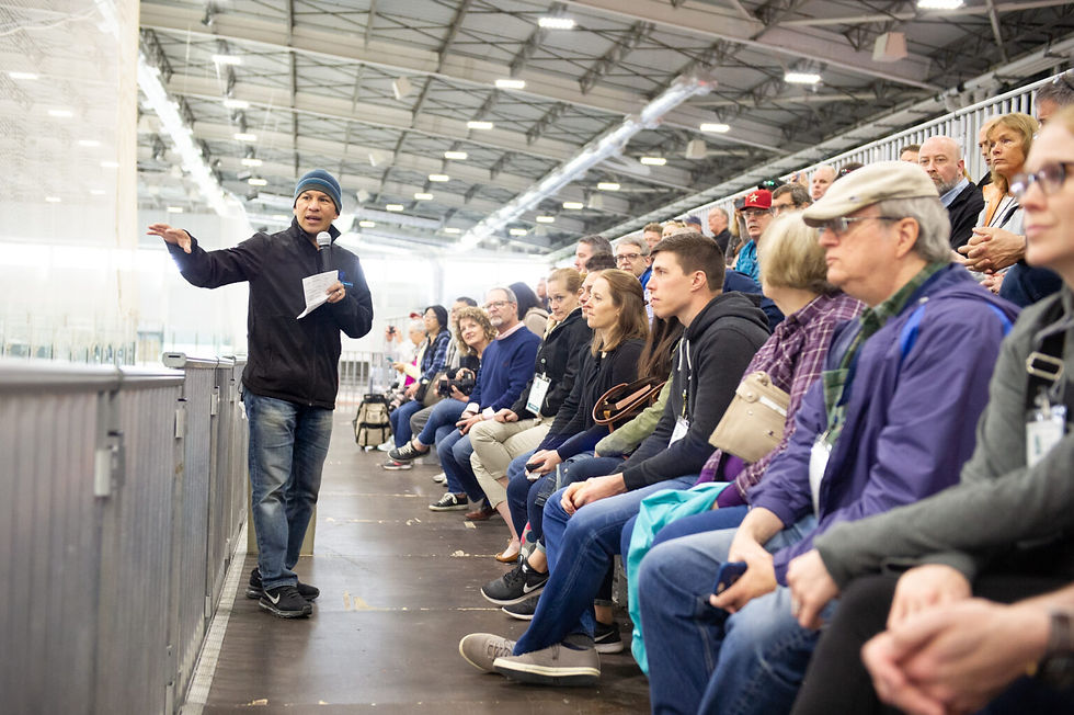 A speaker with a microphone addresses an attentive audience in a large indoor venue. People are seated in rows, listening intently.