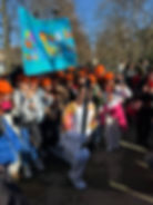 A person in colorful gear kneels holding a torch, surrounded by kids in orange hats. They hold a bright sign in a park setting. Jovial mood.