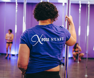 A woman in a purple shirt with the words “Xpose Staff” in white, standing next to a steel dance pole