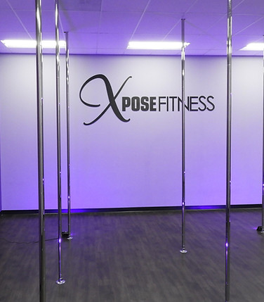 The Xpose Fitness pole dance studio room. Several steel poles sit in front of a wall with large letters reading “Xpose Fitness”