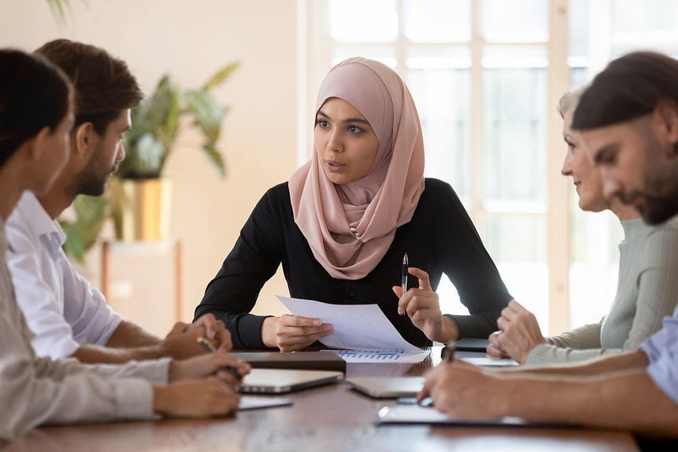 Five people in a meeting room, discussing documents. A woman in a pink hijab leads the conversation. Bright window and plants in background.