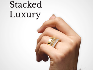 Hand wearing stacked gold and diamond rings with "Stacked Luxury" text; fur sleeve visible against a gray background, symbol above.