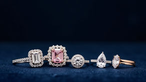 Five elegant rings with different engagement rings are displayed on a dark blue surface. The center ring showcases a pink gemstone.