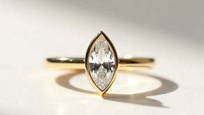 Close-up of a horizontal marquise-cut diamond in a minimalist yellow gold bezel setting on a slender band, casting a long shadow on a white surface.