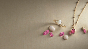 Gold jewelry set with pink and white gemstones on a textured beige background. Includes a necklace, ring, earrings, and loose stones.