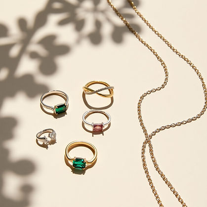 Gold and silver rings with green and pink gemstones, alongside a gold chain on a beige surface. Leaf shadows create a decorative pattern.