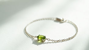 Delicate gold chain bracelet with green peridot gemstone on a white surface. Soft shadows and white flowers in the background, creating a serene mood.