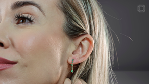 Close-up of a woman showcasing a green emerald gemstone earring with a serene expression. Blonde hair partially covers a gray background.